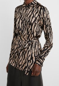 Chemise à manches longues en imprimé animalier noir et beige, avec un col à revers, une fermeture boutonnée et un détail de ceinture nouée. Tissu à texture lisse.
