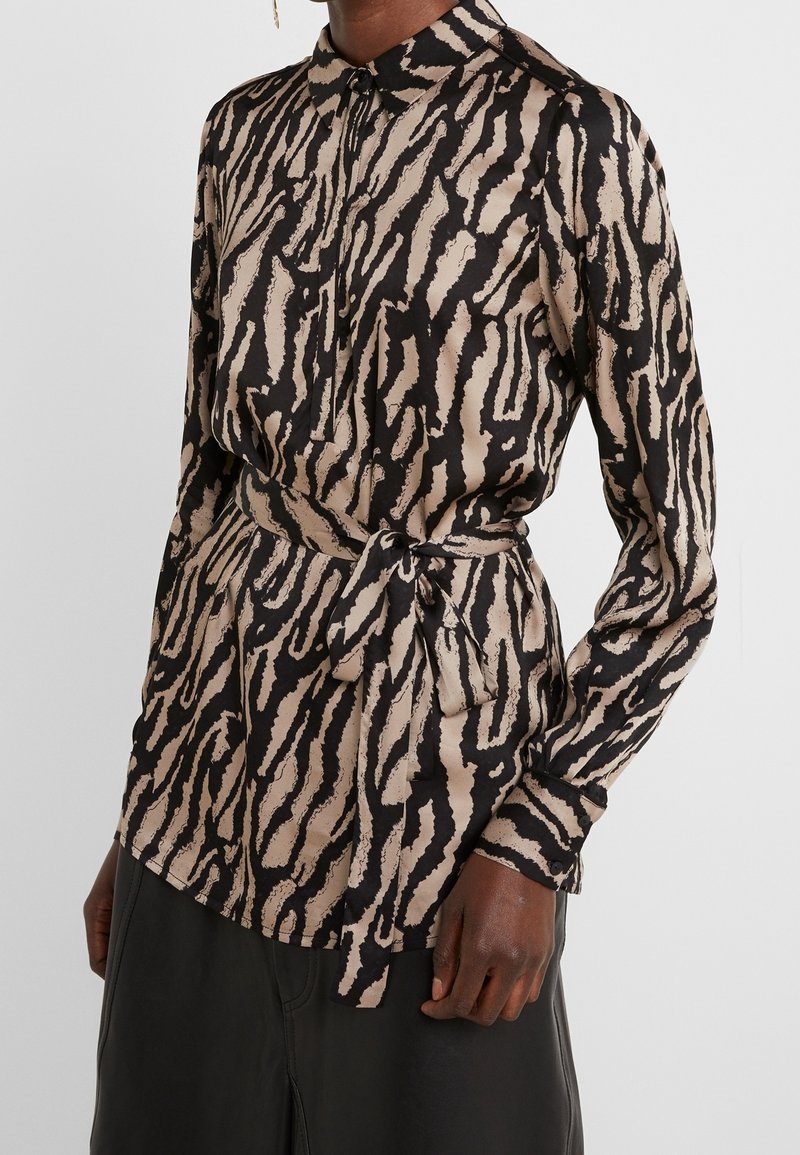 Chemise à manches longues en imprimé animalier noir et beige, avec un col à revers, une fermeture boutonnée et un détail de ceinture nouée. Tissu à texture lisse.