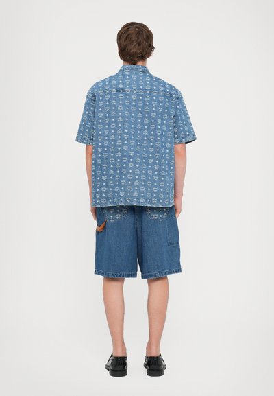 Ljusblå denimskjorta med kort ärm och upprepat vitt grafiskt mönster, tillsammans med knälånga denimshorts. Slät textur, avslappnad passform.