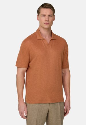 Uomo che indossa una polo a maniche corte di colore ruggine e pantaloni beige, in piedi davanti a uno sfondo chiaro e uniforme.