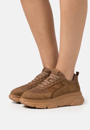 Baskets en daim marron et mesh avec semelles épaisses en caoutchouc portées pieds nus sur un fond blanc uni.