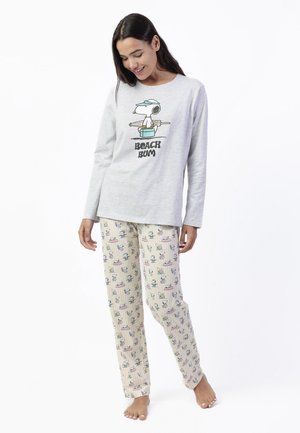 Mujer con una camiseta de manga larga gris con un gráfico de Snoopy "Beach Bum" y pantalones de pijama de color claro con patrones de Snoopy, de pie descalza.