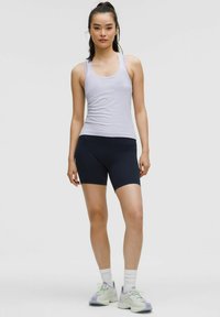 Haut sans manches violet clair, shorts bleu marine foncé et chaussures de sport vert clair avec des accents violets, sur un fond gris uni.