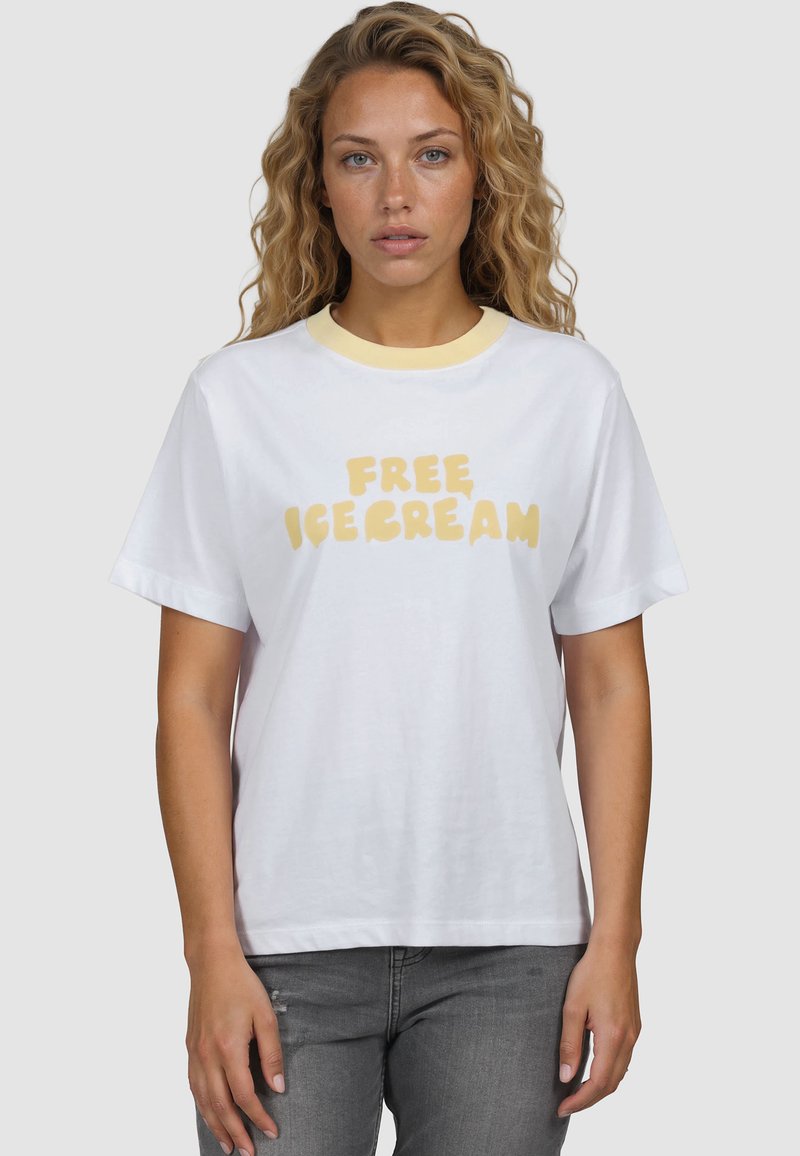 Γυναίκα με σγουρά μαλλιά που φοράει λευκό T-shirt με κίτρινο γιακά και την επιγραφή "ΔΩΡΕΑΝ ΠΑΓΩΤΟ", στέκεται μπροστά σε απλό φωτεινό φόντο.