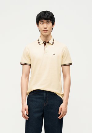 Giovane uomo che indossa una polo beige con colletto e maniche a strisce scure, abbinata a jeans blu scuro, in piedi davanti a uno sfondo semplice.