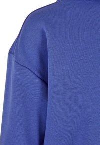 Detailní záběr na modrou mikinu, která má hladkou texturu, ribbený límec a strukturované ramenní švy. Látka vypadá měkká a odolná.