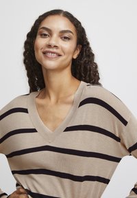 Beige Pullover mit dunklen Streifen, V-Ausschnitt, weicher Textur, lockerer Passform und langen Ärmeln. Model präsentiert die Vorderansicht und lächelt.