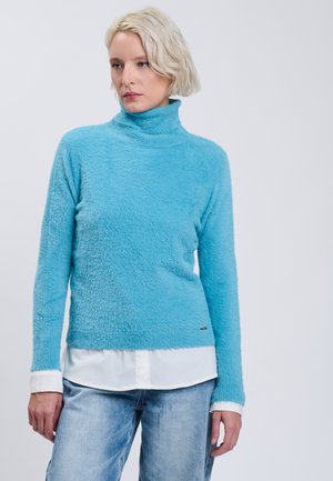 Mujer rubia con un jersey de cuello alto azul claro y peludo encima de una camisa blanca y vaqueros azules, de pie contra un fondo liso y claro.