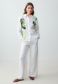 Camicia bianca in lino con motivi a foglia verde sul davanti, abbinata a pantaloni larghi bianchi in lino e sandali piatti marroni con cinturini.