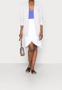 Vaaleanharmaa bleiseri purppuranvärisen topin päällä, yhdistetty valkoiseen kietaisuhameeseen. Mustat liukuvaikutteiset sandaalit ja pieni ruskea olkalaukku täydentävät asukokonaisuuden.