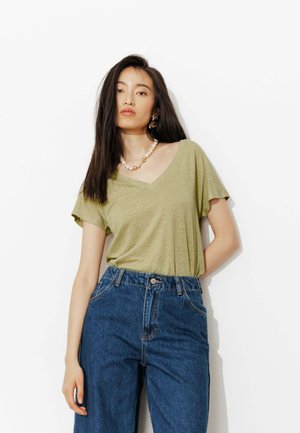 Jeune femme aux longs cheveux noirs portant un t-shirt vert olive à col en V, un jean bleu taille haute, un collier de perles et des boucles d'oreilles en or, debout contre un mur blanc.