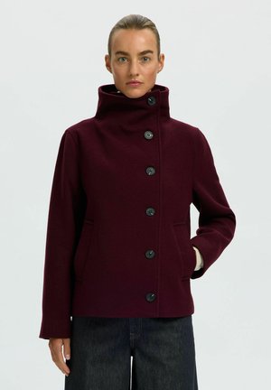Femme aux cheveux tirés en arrière portant un manteau boutonné bordeaux foncé à col montant et un pantalon foncé, debout devant un fond uni.