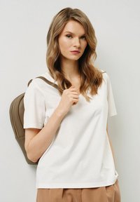 Biała bawełniana koszulka z dekoltem w serek, krótkimi rękawami i luźnym krojem. Model trzyma brązowy plecak z zamkiem błyskawicznym.