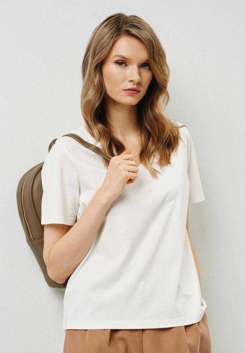 Biała bawełniana koszulka z dekoltem w serek, krótkimi rękawami i luźnym krojem. Model trzyma brązowy plecak z zamkiem błyskawicznym.