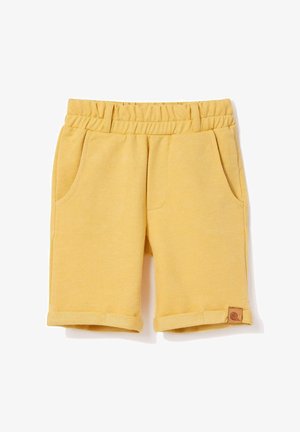 Uśmiechnięty chłopiec stojący z ręką w kieszeni, ubrany w granatową koszulkę polo, żółte szorty i beżowe mokasyny na jednolitym jasnym tle.