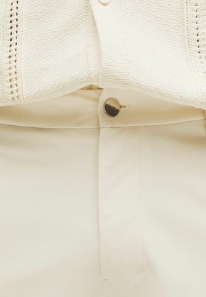 Cardigan en tricot couleur crème sur un pantalon crème taille haute et ajusté. Comprend un bouton en métal visible et des détails cousus sur la taille.