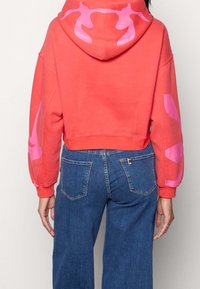 Sudadera corta de color coral con diseños abstractos en rosa en las mangas y la capucha, hecha de material suave. Combinada con jeans de mezclilla azul.