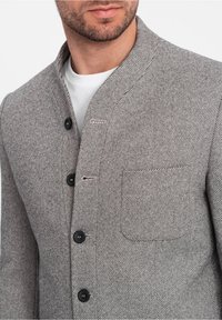 Uomo che indossa una giacca a righe diagonali grigia con bottoni neri e una tasca sul petto sopra una maglietta bianca a girocollo.