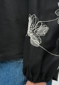 Čierna blúzka s dlhými, nafúknutými rukávmi, zdobená zložitou bielou kvetinovou výšivkou na rukáve, vyrobená z ľahkej tkaniny.