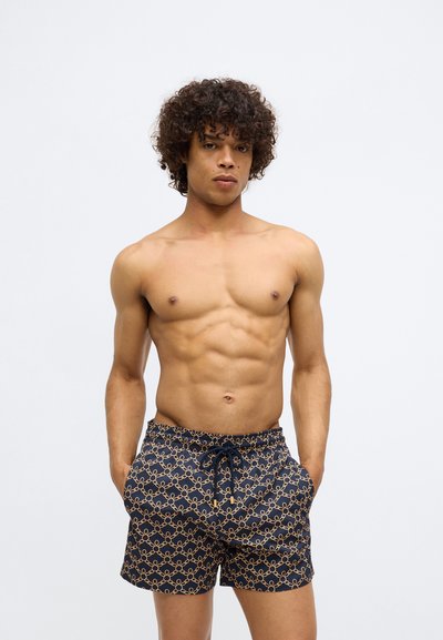 Jauns vīrietis ar cirtainiem matiem, ģērbies tumši zilās peldbiksēs ar bežīgu ķēdes rakstu, stāv ar rokām kabatās uz vienkārša fona.