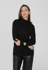 Schwarzer Rollkragenpullover aus weichem, geripptem Stoff mit seitlichen Schlitzen, kombiniert mit glänzenden schwarzen Hosen und goldenen Armreifen.