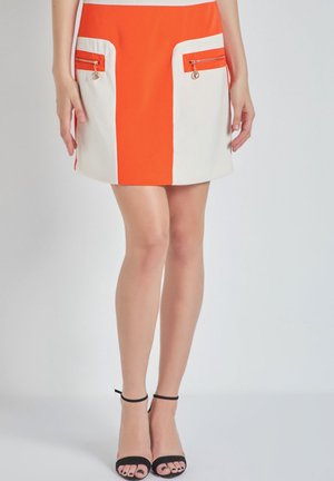 Mujer que lleva una falda corta naranja y blanca con bolsillos con cremallera y sandalias de tacón alto con correa en el tobillo negras, de pie contra un fondo liso.