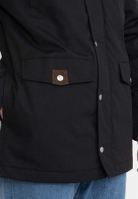 Giacca nera con chiusura frontale a bottoni, due tasche laterali con accenti marroni e una consistenza del tessuto liscia e resistente.