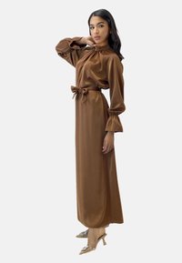 Abito maxi in raso marrone con vita legata, colletto alto e maniche a sbuffo. Indossato con scarpe con tacco trasparente decorate con dettagli.