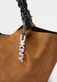 Bolsa de camurça castanha com uma alça preta trançada, apresentando um destaque prateado com o logótipo DKNY. O interior parece escuro e sem adornos.