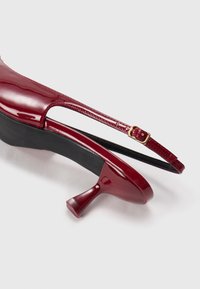 Punainen lakattu nahka slingback-kengä, jossa on kaareva korko, säädettävä hihna kultaisella soljella ja smooth, kiiltävä pinta.