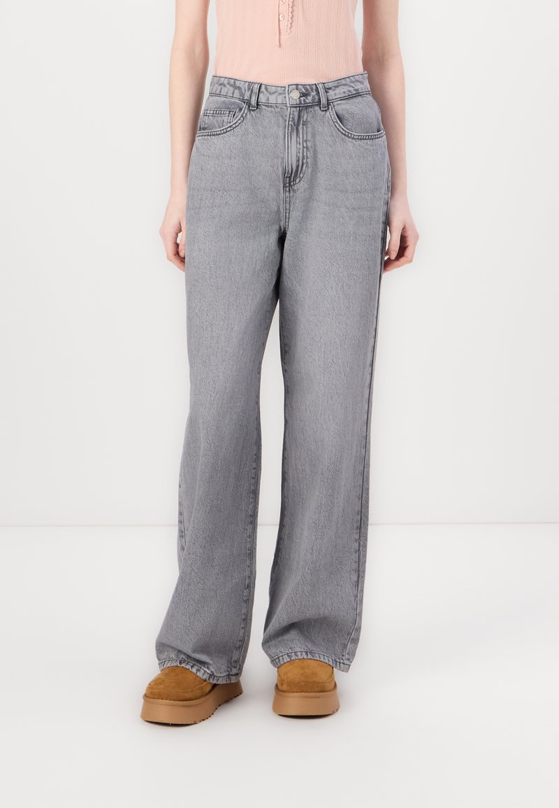 Jeans de corte ancho en tonos grises con un efecto de lavado desgastado, diseño de cinco bolsillos, cierre de botón metálico y detalles de costura visibles, combinados con calzado beige.