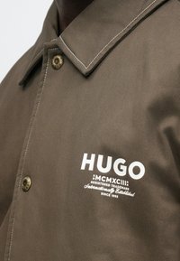 Καφέ σακάκι με γιακά, με λευκή ραφή και μεταλλικά κουμπιά, με το λογότυπο "HUGO" και κείμενο στο στήθος.