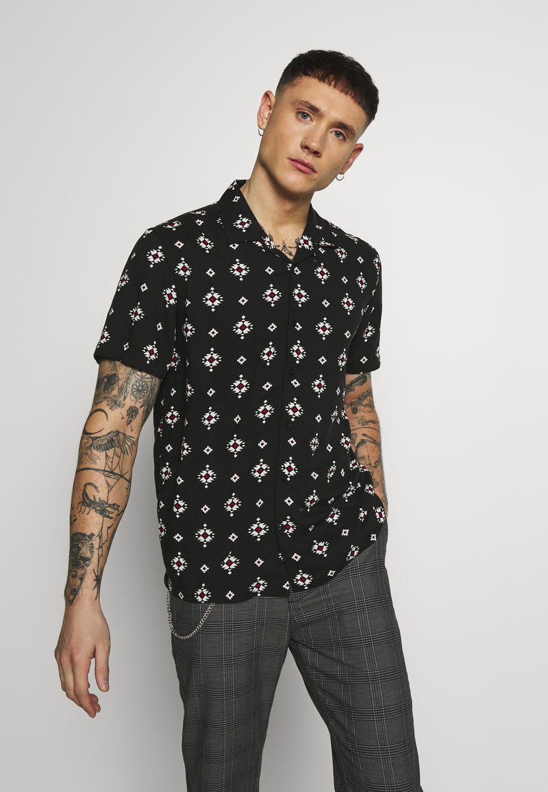 Zwart shirt met korte mouwen, voorzien van een geometrisch patroon in wit en rood, knopen aan de voorkant, in relaxed fit, gecombineerd met grijs geruite broek.