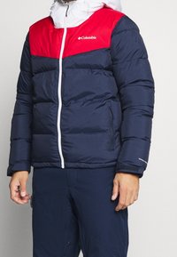 Columbia Daunenjacke mit navyfarbenem Körper, rotem oberen Bereich, weißem Reißverschluss und verstellbarem Kapuze. Verfügt über Reißverschlusstaschen und Klettverschlussmanschetten.