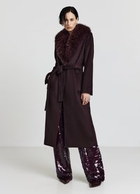 Cappotto lungo di un elegante color bordeaux scuro con colletto in pelliccia sintetica, vita con cintura e due tasche frontali, indossato sopra pantaloni con paillettes. Texture liscia, design elegante.