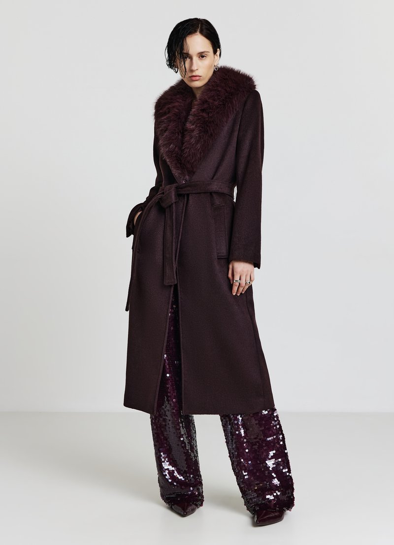 Cappotto lungo di un elegante color bordeaux scuro con colletto in pelliccia sintetica, vita con cintura e due tasche frontali, indossato sopra pantaloni con paillettes. Texture liscia, design elegante.