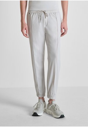 Hombre con pantalones jogger gris claro con cordón y zapatillas a juego, de pie contra un fondo liso y claro.