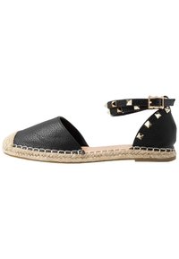 Sandalia plana de espadrille de cuero negro con suela de yute trenzado, tachuelas doradas en forma de pirámide y dobles correas al tobillo con cierre de hebilla.
