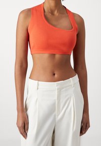 Kobieta ubrana w asymetryczny pomarańczowy prążkowany crop top i białe, plisowane spodnie z wysokim stanem, stojąca z luźno opadającymi ramionami.