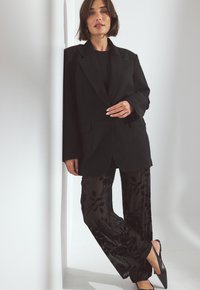 Črn prevelik blazer z zavihki, vparen s prosojnimi črnimi hlačami s cvetličnim vzorcem in črnimi pointešnimi čevlji. Gladka textura.