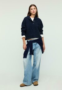 Navy blue knit sweater with a high collar and button detail, paired with light blue flared jeans and tan suede shoes. Long straight hair.