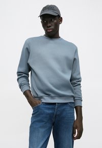 Lyseblå crewneck-genser laget av mykt stoff, kombinert med blå jeans. En grå caps med brodert tekst fullfører antrekket.