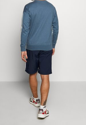 Blauer Sweatshirt mit Rundhalsausschnitt und langen Ärmeln, kombiniert mit marineblauen Shorts. Weiße Akzente auf den Schultern. Sneaker mit roten und grauen Details.