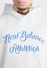 Hombre con sudadera gris claro con el texto azul "New Balance Athletics" en el pecho, mostrando parte de la cara y el cuello.