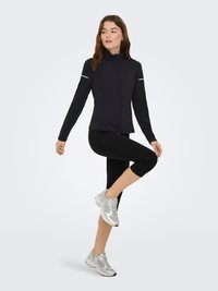 Zwarte sportieve jas met een hoge kraag, ritssluiting en reflecterende accenten, gecombineerd met zwarte capri-legging en zilveren hardloopschoenen.