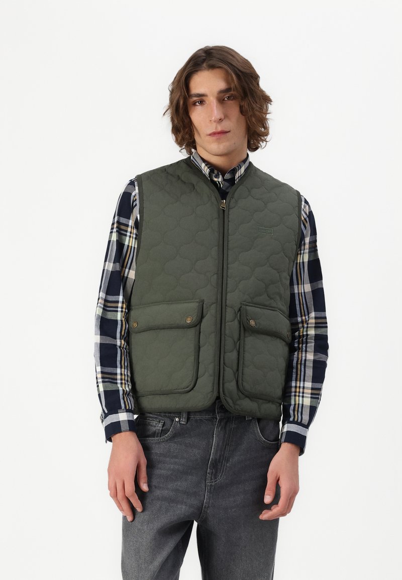 Quilted green vest with a high collar, zip closure, and two front pockets; worn over a plaid long-sleeve shirt and gray jeans.