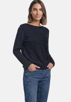 Femme aux cheveux mi-longs portant un t-shirt noir à manches longues et un jean bleu, debout, les mains dans les poches, sur fond blanc.