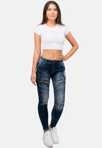 Jeans skinny in denim blu scuro con dettagli in zip e strappi, abbinati a una t-shirt cropped bianca e sneakers bianche.
