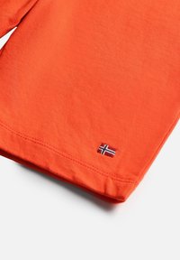 Tissu orange à manches longues avec des coutures visibles près de l'ourlet et un petit drapeau norvégien brodé près du bord inférieur.