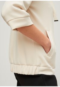 Lichtbeige pullover met een ronde halslijn, ribbelmanchetten en een gathered zoom. Beschikt over een zijzak met een slanke opening.